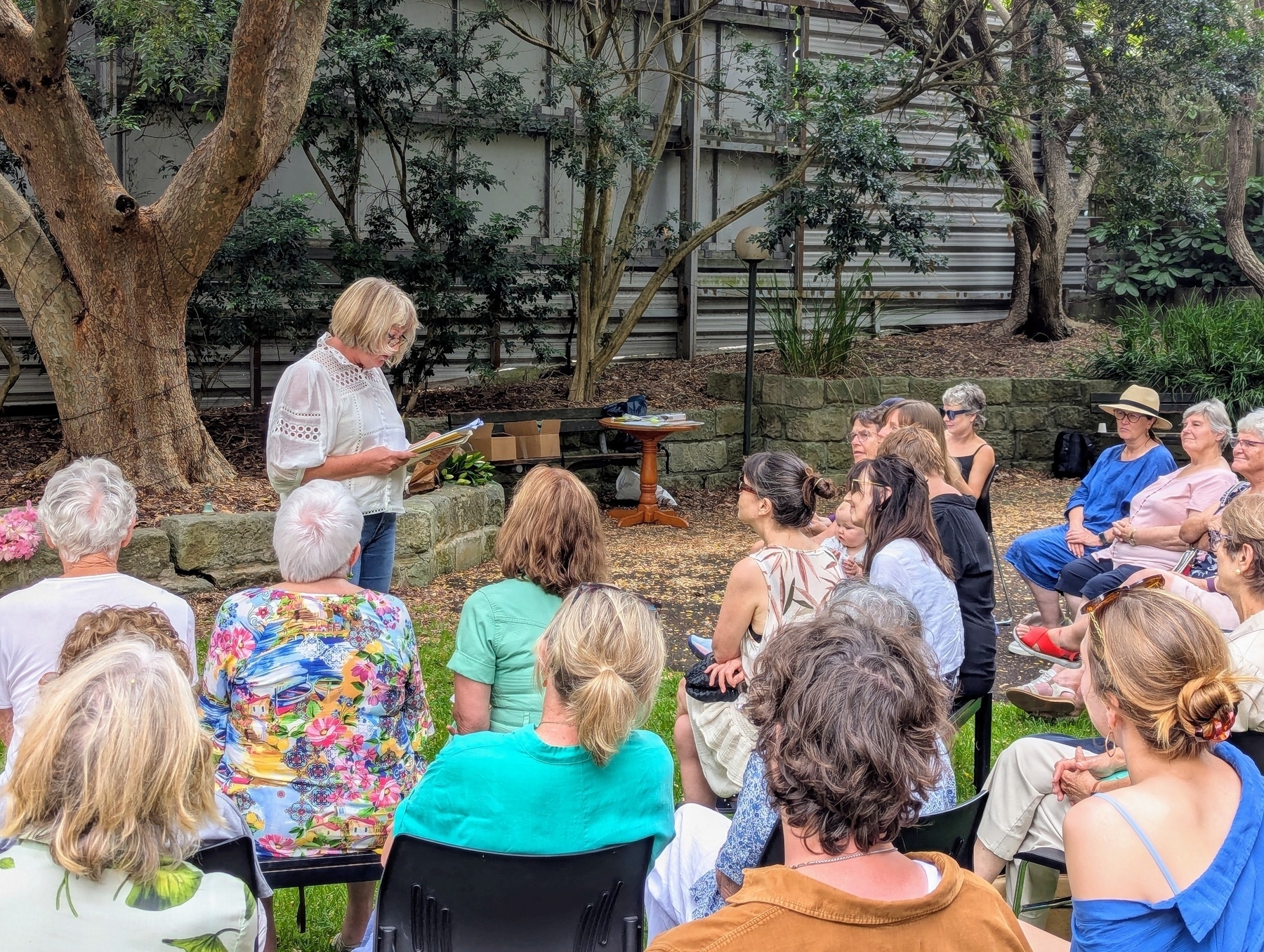 A woman is speaking to a seated outdoor audience in a garden setting.