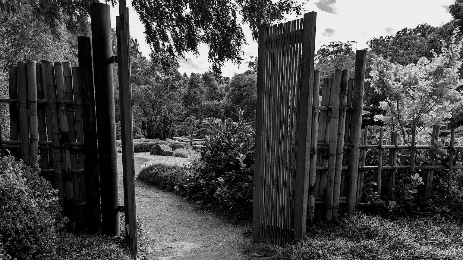 A rustic bamboo gate opens to a lush, tranquil Japanese-style garden path surrounded by trees and shrubs.